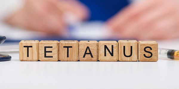 Holzwürfel mit schwarzen Buchstaben bilden das Wort TETANUS auf einer weißen Fläche. Rechts liegt eine Spritze, im Hintergrund unscharfe Hände. Darstellung weist auf Impfberatung oder Vorsorge hin.