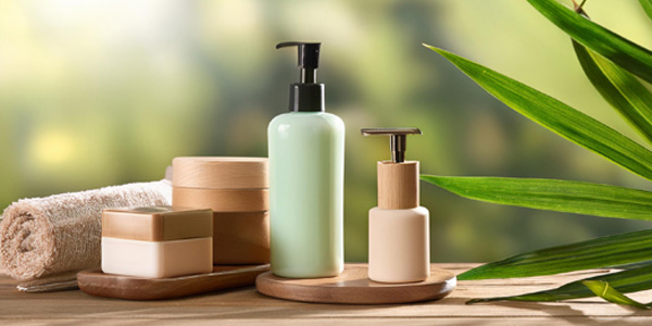 Auf einem Holztisch stehen zwei Spenderflaschen für Pflegeprodukte sowie mehrere Kosmetikdosen. Daneben liegt ein gerolltes Handtuch, rechts sind grüne Blätter zu sehen, Hintergrund unscharf.
